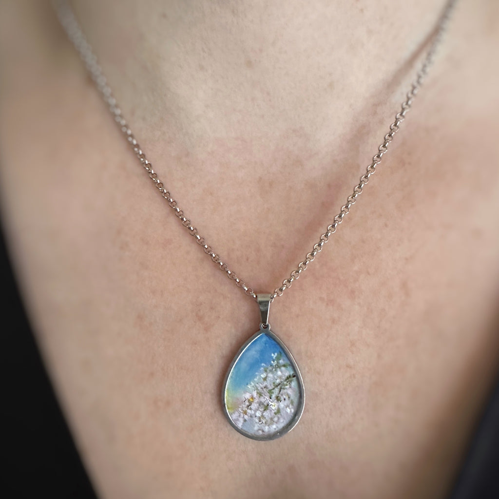Silver colored tear drop shaped pendant, hand painted, depicting a branch of cherry blossoms against a sunset sky. Shown on model.