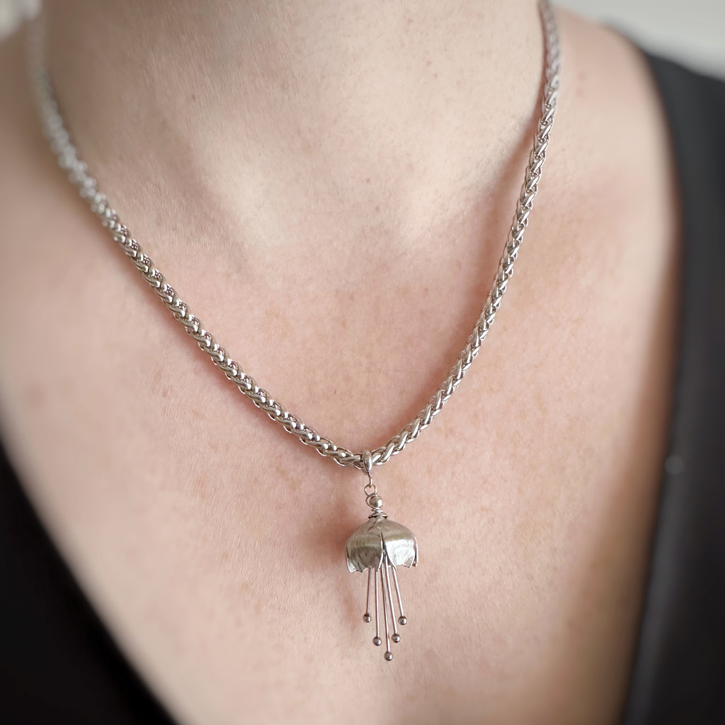 Hand made and scuplted surgical grade steel inverted flower pendant with a satin finish with long dangling stamen. Hanging from a thick chain necklace. Shown on model.