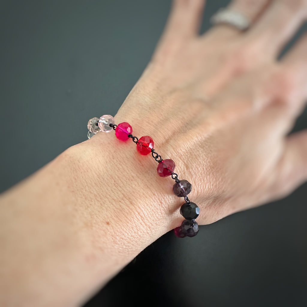 pink and red spectrum faceted glass beaded bracelet ,with hand wrapped, secure links, displayed on a table. Shown worn on wrist.