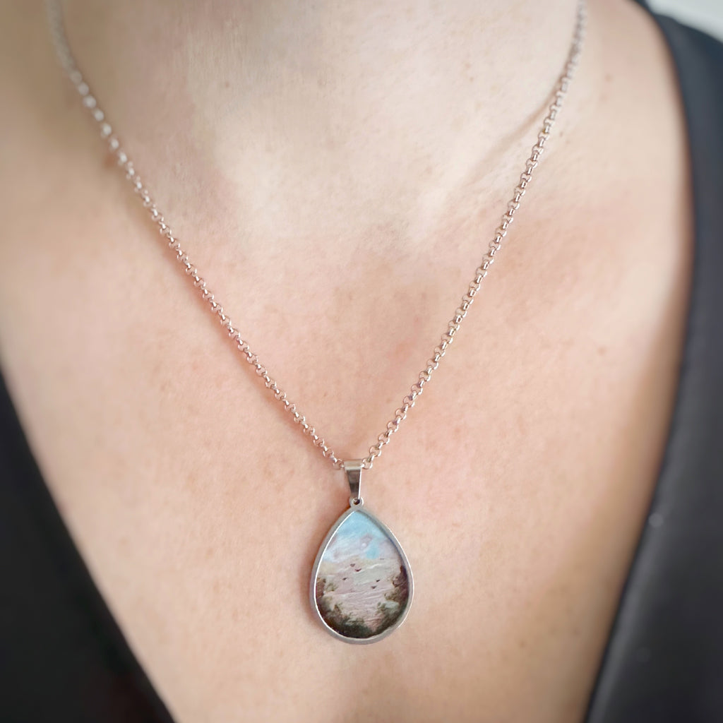 Silver color teardrop shaped pendant, hand painted depicting a flock of birds taking flight at sunset, near the edge of a hedgerow. Shown on model.