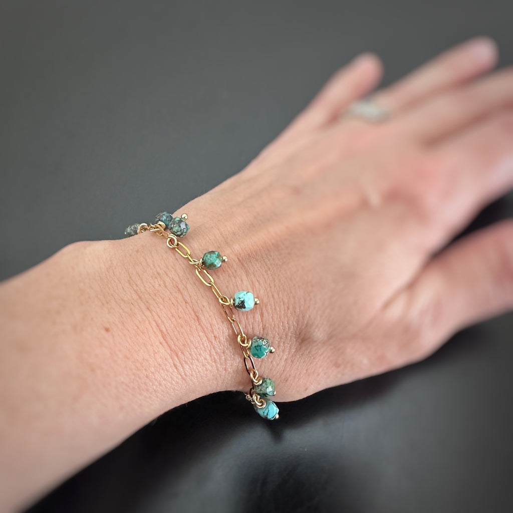 Faceted cube shaped turquoise beads, dangling like fringe off of a gold-tone paperclip style chain bracelet. Shown on model.