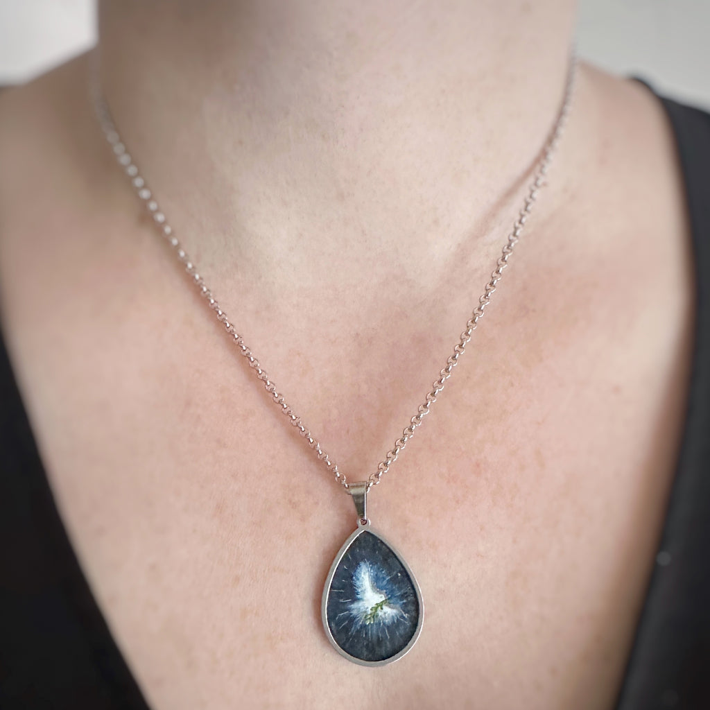 Silver color teardrop shaped pendant, hand painted depicting a white dove carrying an olive brance and set against a midnight blue sky. Shown on model.