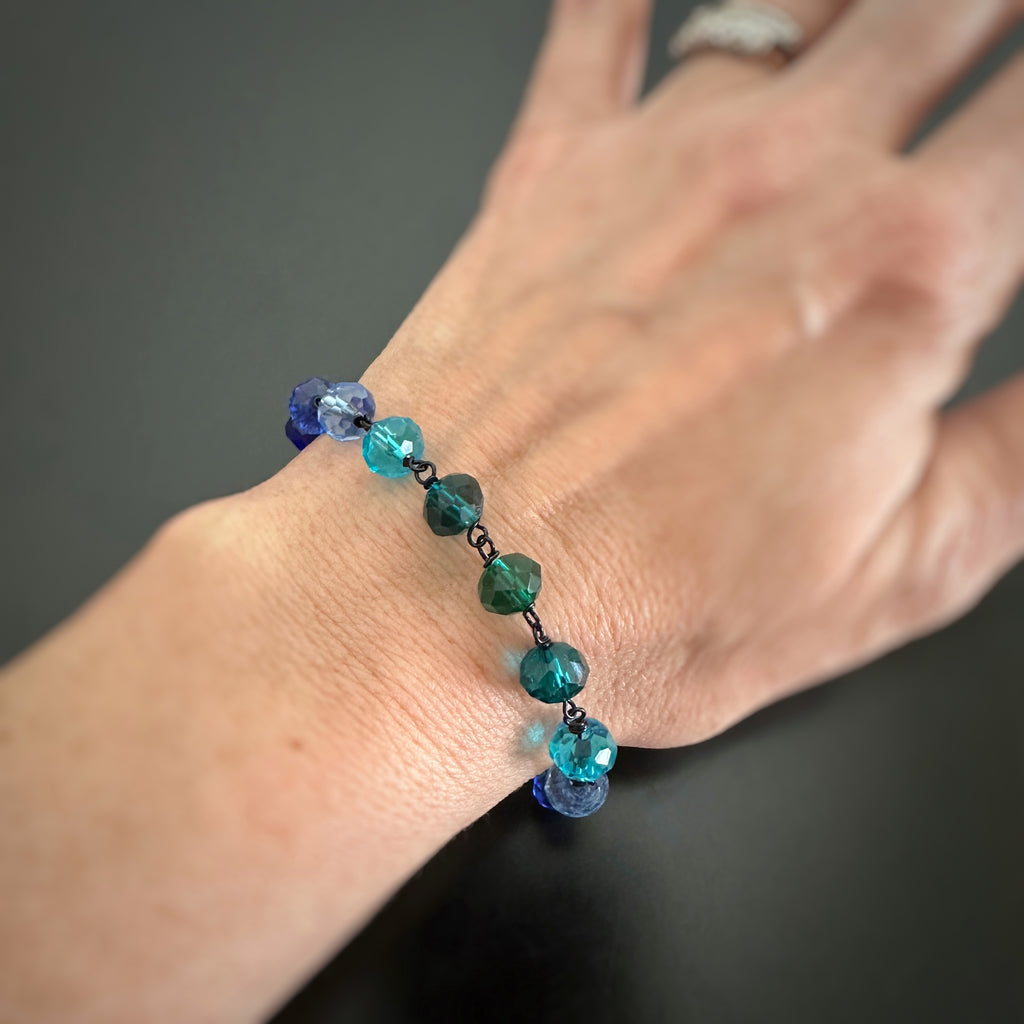 Blue and aqua spectrum faceted glass beaded bracelet ,with hand wrapped, secure links, displayed on a table. Shown worn on wrist.