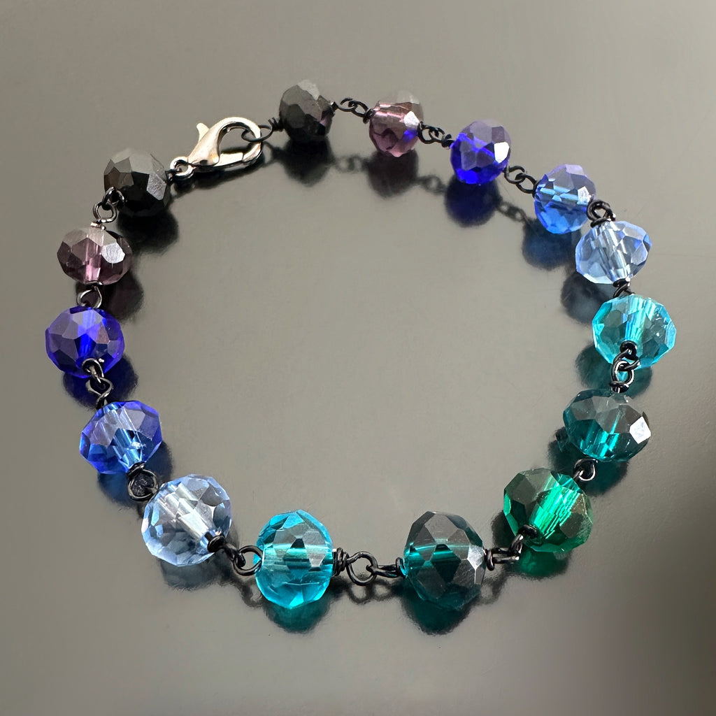Blue and aqua spectrum faceted glass beaded bracelet ,with hand wrapped, secure links, displayed on a table.