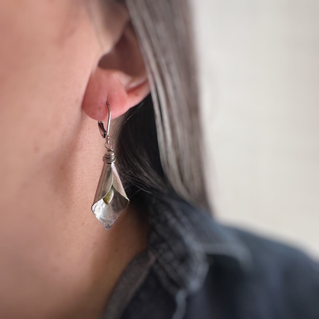 Hand formed silver tone surgical steel inverted calla lily drop earrings with pale green Czech glass stamen. The earrings use lever-back style ear wires. Shown and worn.