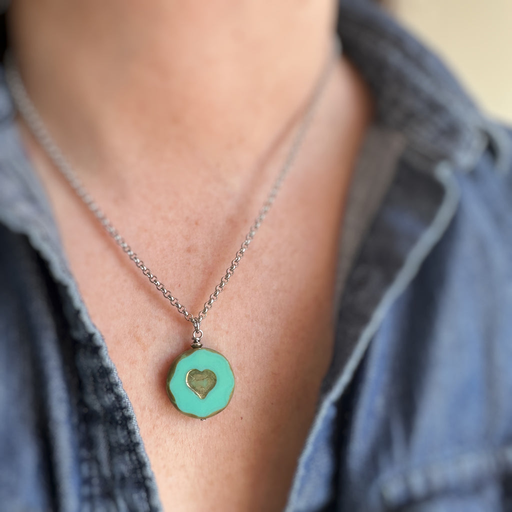 Pendant necklace with turquoise glass coin shaped bead with crackled golden hearts in the center. The surgical steel chain is a rolo-style link.