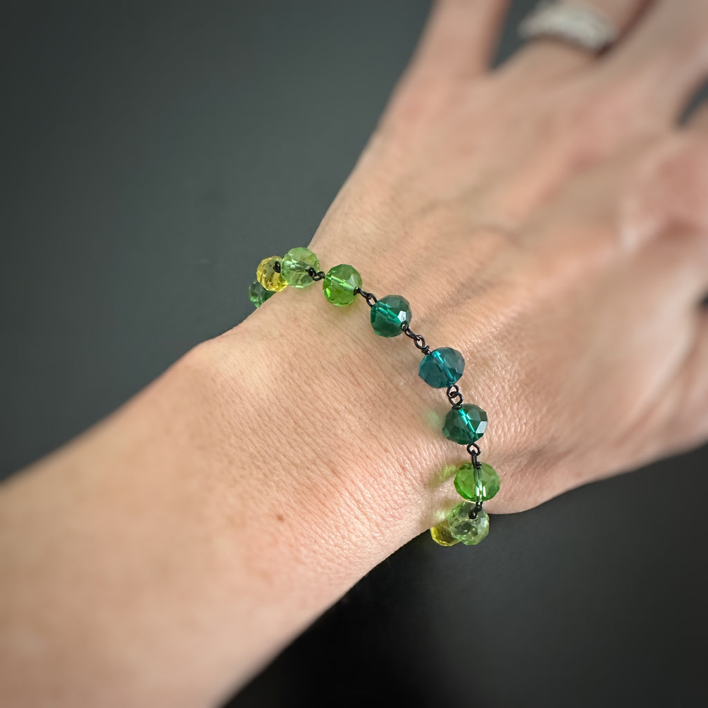 Green and yellow spectrum faceted glass beaded bracelet ,with hand wrapped, secure links, displayed on a table. shown worn on wrist