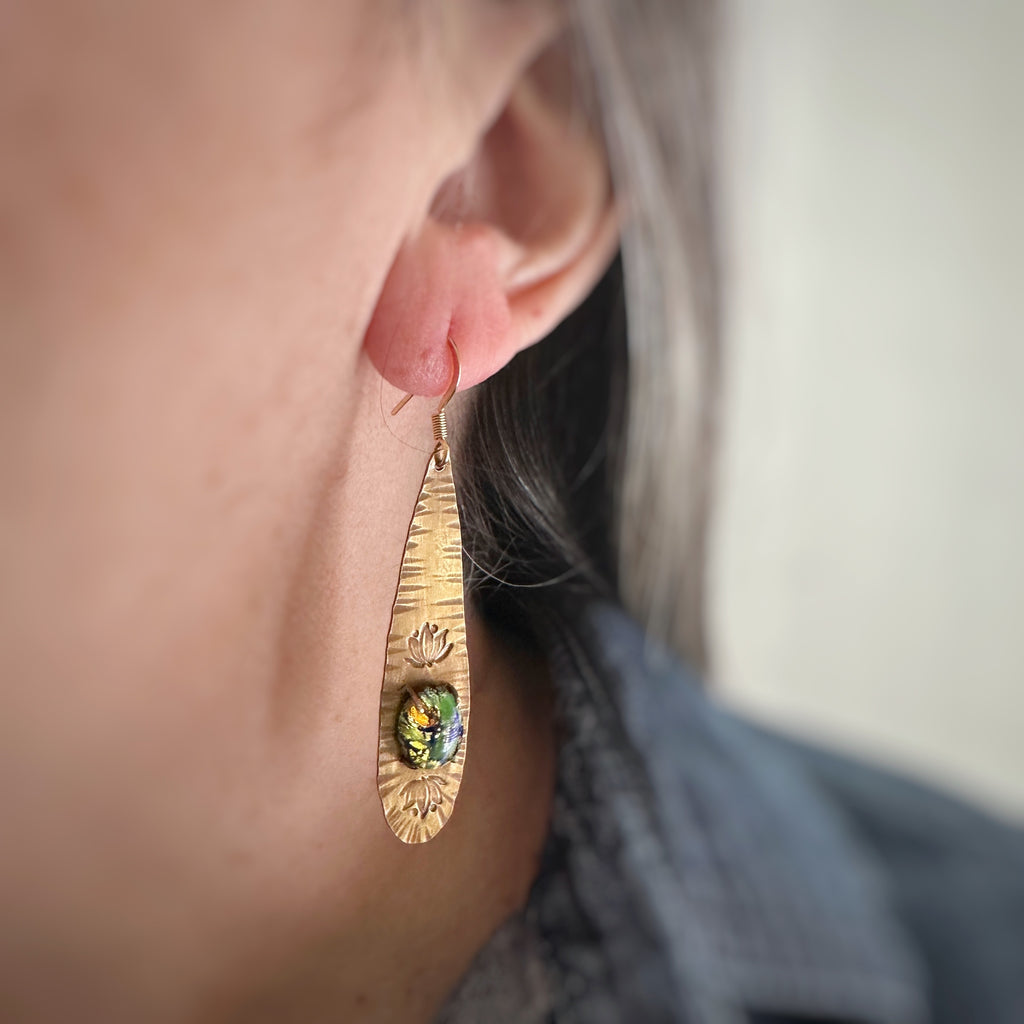 long brass teardrop earrings with french hook ear wires. The tear drop has hammer marks and stamped lotus flowers with a focal of a green opal glass oval, prong-set near the bottom. shown as worn