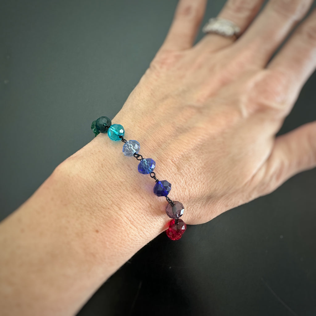 A collection of rainbow spectrum faceted glass beaded bracelets in various colors, with hand wrapped, secure links, displayed on a table. shown worn on wrist