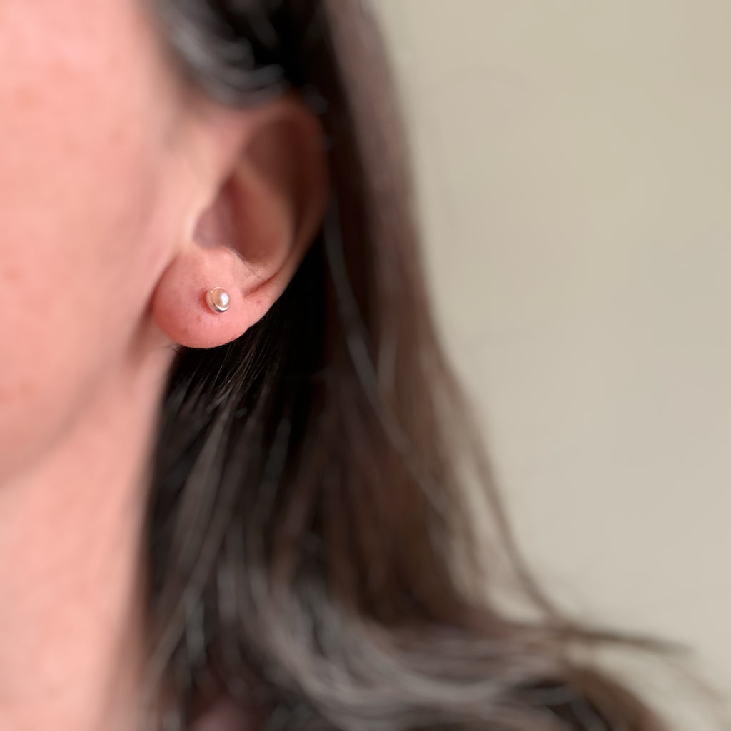 Tiny round peachy pink freshwater pearl stud earrings made with sterling silver shown on model.