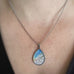 Silver colored tear drop shaped pendant, hand painted, depicting a branch of cherry blossoms against a sunset sky. Shown on model.