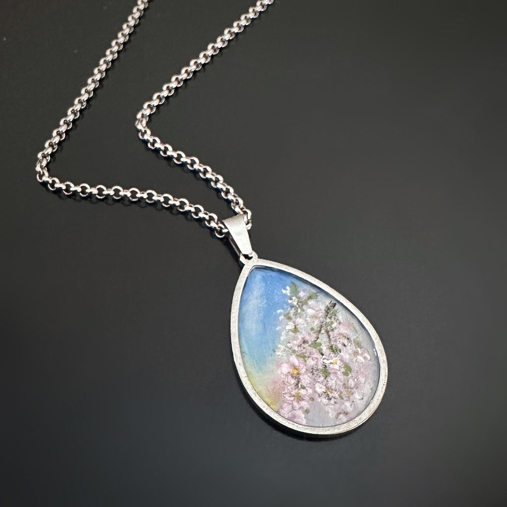 Silver colored tear drop shaped pendant, hand painted, depicting a branch of cherry blossoms against a sunset sky.