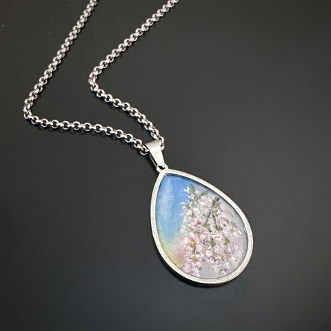 Silver colored tear drop shaped pendant, hand painted, depicting a branch of cherry blossoms against a sunset sky.