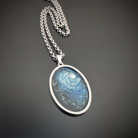Silver color oval shaped pendant, hand painted depicting a full moon in a dark but hazy sky, and a row of trees on the left side.