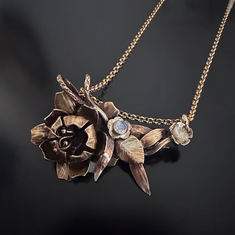 Golden brass and surgical steel pendant necklace, evoking a leaf filled bouquet highlighting a peony and a tiny wild rose with a small moonstone and hanging asymmetrically from a rolo style chain.
