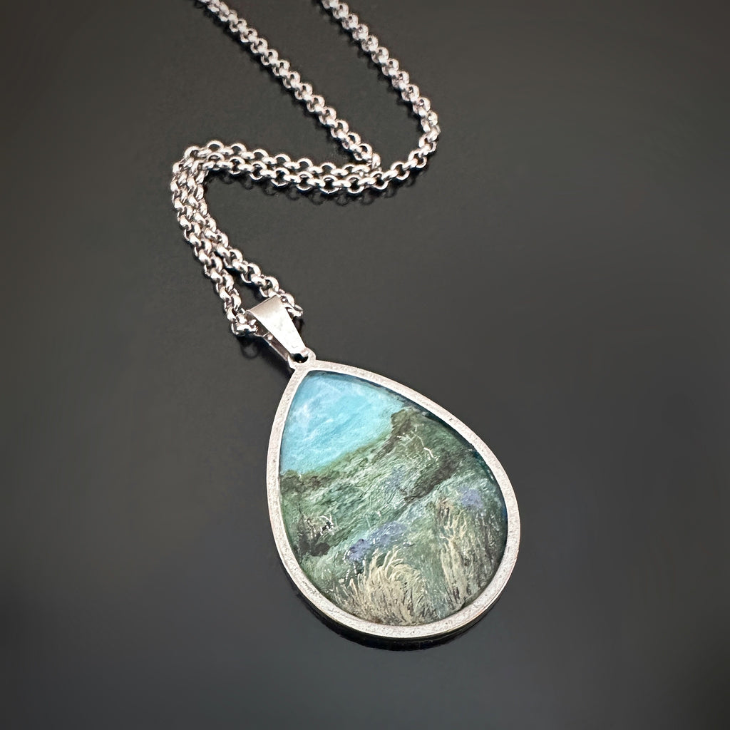 Silver color teardrop shaped pendant, hand painted depicting a summer field filled with tall grasses, brush, and wildflowers.