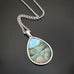 Silver color teardrop shaped pendant, hand painted depicting a summer field filled with tall grasses, brush, and wildflowers.