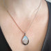 Silver color teardrop shaped pendant, hand painted depicting a flock of birds taking flight at sunset, near the edge of a hedgerow. Shown on model.