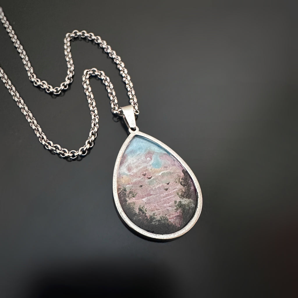 Silver color teardrop shaped pendant, hand painted depicting a flock of birds taking flight at sunset, near the edge of a hedgerow.