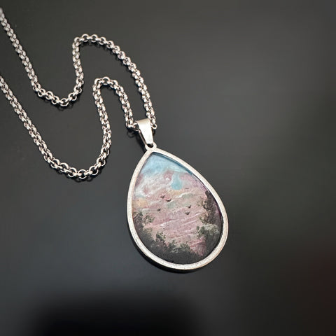 Silver color teardrop shaped pendant, hand painted depicting a flock of birds taking flight at sunset, near the edge of a hedgerow.