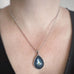 Silver color teardrop shaped pendant, hand painted depicting a white dove carrying an olive brance and set against a midnight blue sky. Shown on model.