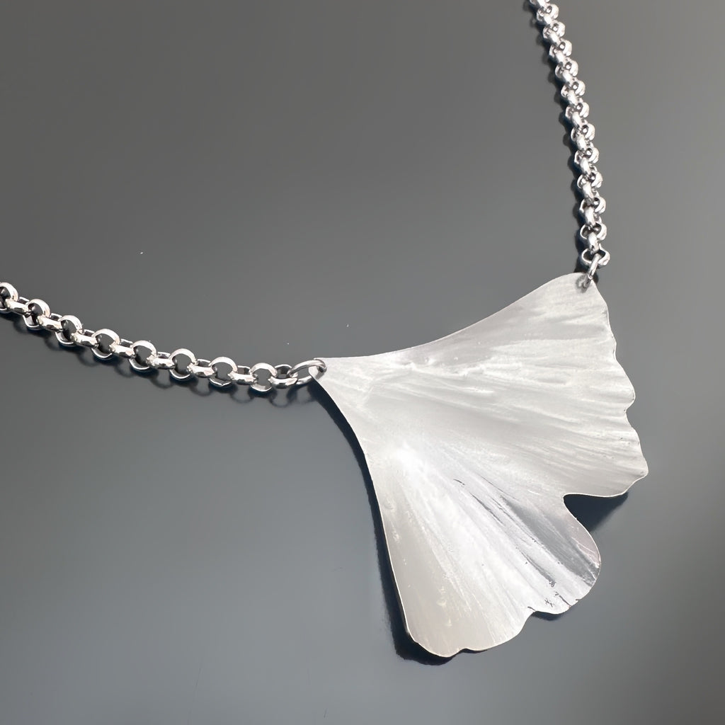 Silver tone surgical steel ginkgo leaf shaped pendant on a rolo style chain against a dark gray background