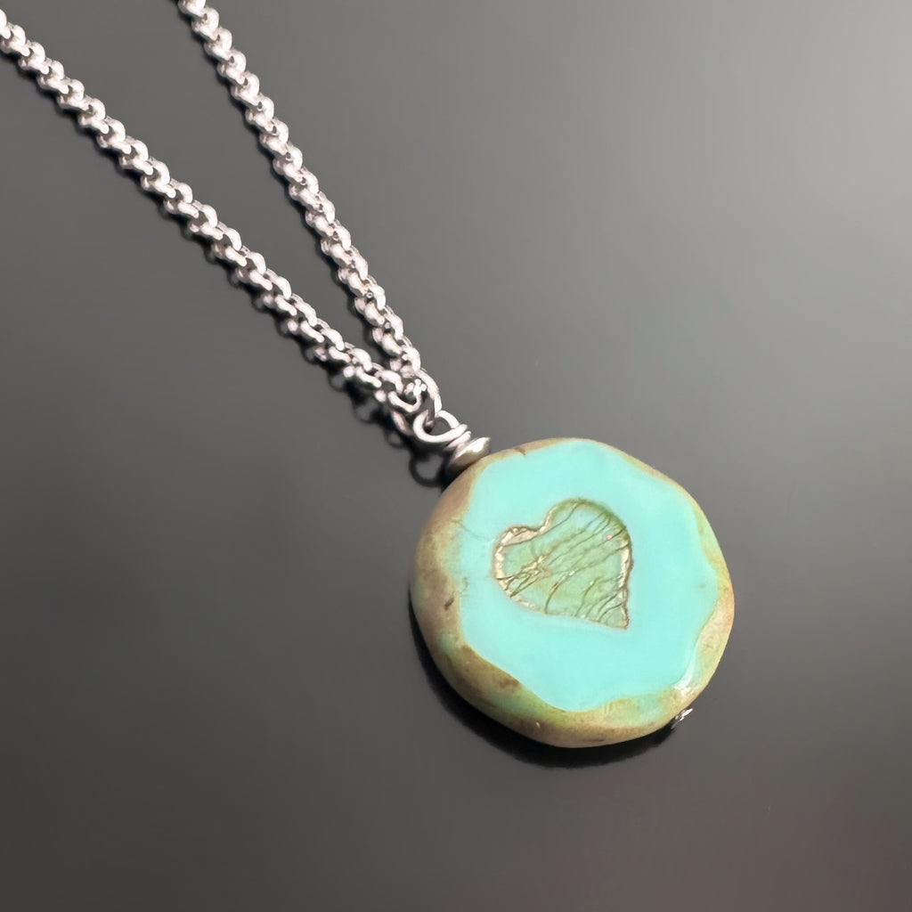 pendant style necklace featuring a turquoise color glass coin shaped bead with crackled golden hearts in the center. The surgical steel chain is a rolo-style link.