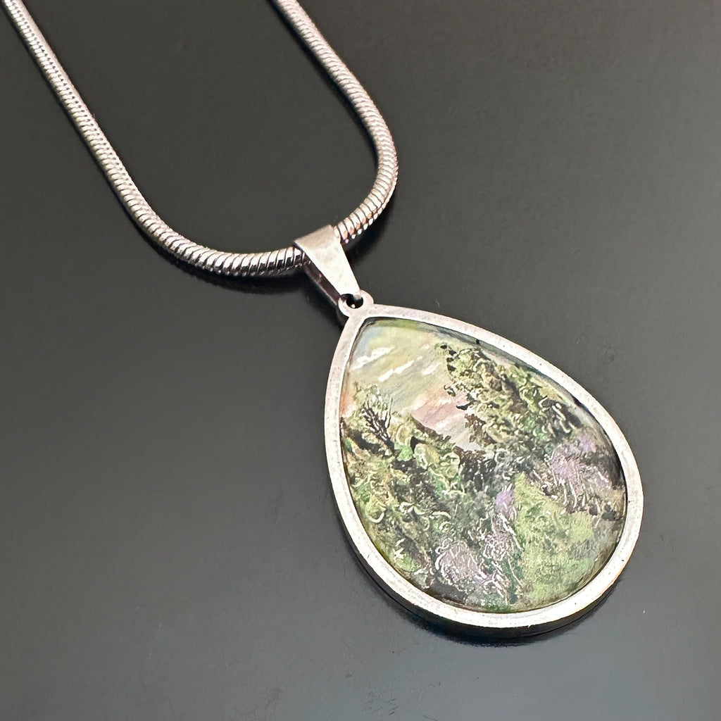 teardrop pendant with a hand painted scenery of a purple thistle lined trail through a pine forest. It hangs from a heavy silver snake style chain.