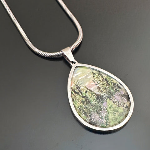 teardrop pendant with a hand painted scenery of a purple thistle lined trail through a pine forest. It hangs from a heavy silver snake style chain.
